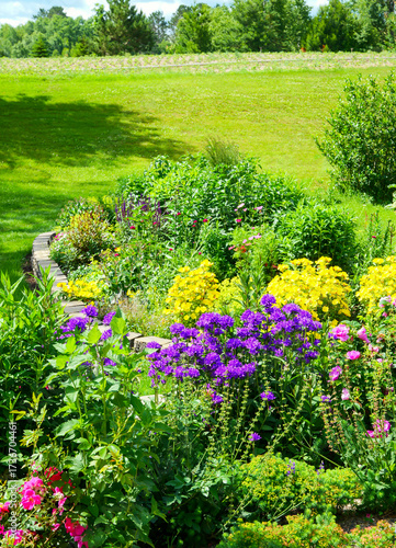 A colorful flower garden, with green lawn, field and trees on a sunny summer day in rural Minnesota.