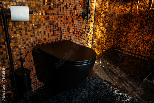 Contemporary bathroom design with orange and gold mosaic tile walls, black toilet and fixtures, dark marble flooring, and a glass shower enclosure.
