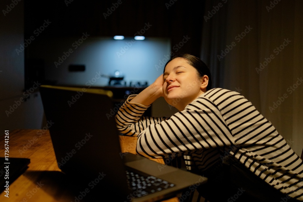 Fototapeta premium A woman sitting at a table with a laptop. She is wearing a striped shirt and has her hair tied back. The background shows a kitchen area with soft lighting.