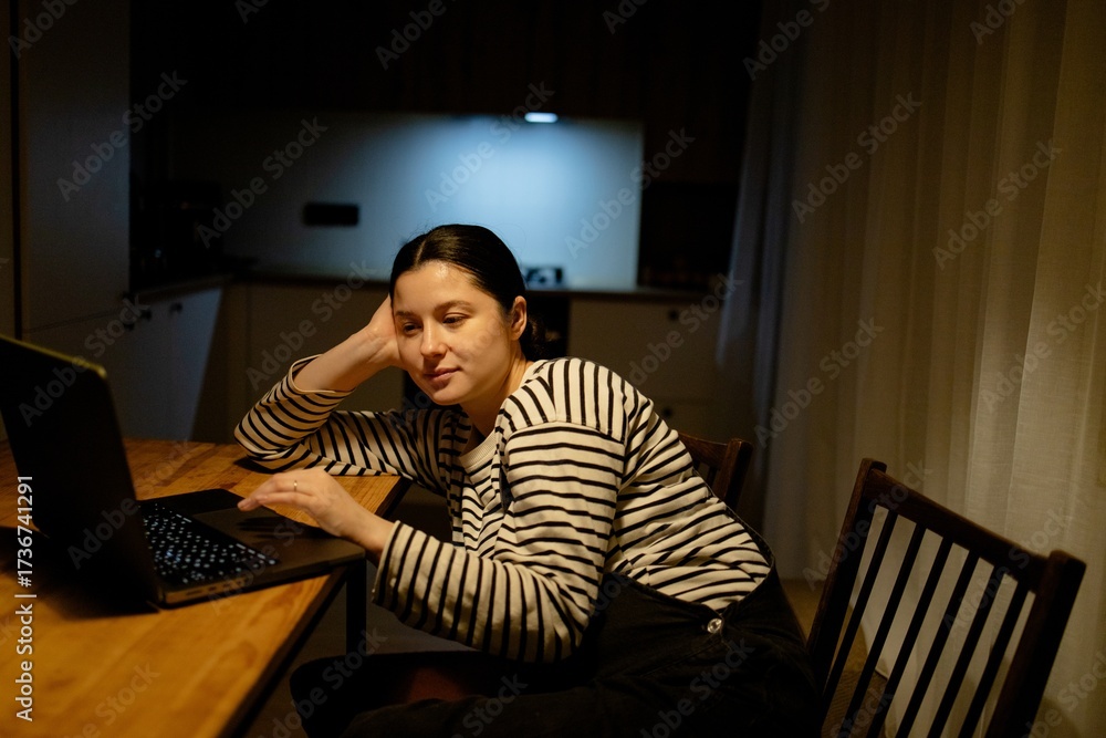 Fototapeta premium A woman sitting at a table with a laptop. She is wearing a striped shirt and has her hair tied back. The background shows a kitchen area with soft lighting.