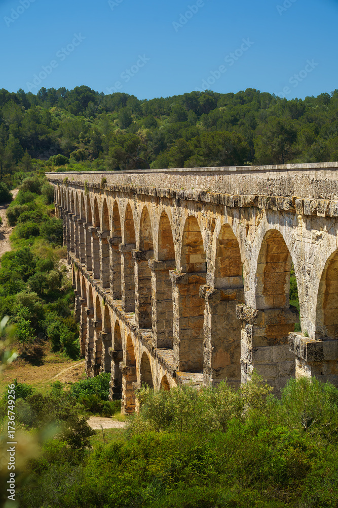 Obraz premium Aqüeducte de les Ferreres, a roman aqueduct, spanning Barranc de les Arcs near Tarragona, sunlit limestone arches and clear blue sky, ancient engineering heritage; ideal for travel ads