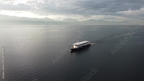Aerial shot of a sleek white ferry gliding across Lake Geneva's glassy, dark waters at dusk. Misty, snow-capped Alps rise in the hazy background under brooding clouds. Serene and majestic.
