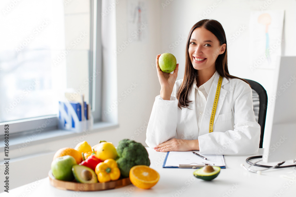 Obraz premium Portrait of happy woman dietitian holding apple, sitting at desk in clinic office with plate of fruits and vegetables, recommending healthy eating