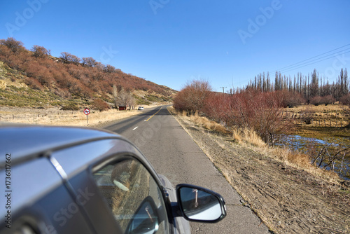 Traveling by car: in the foreground, the side of a vehicle; in the background, the road stretching toward the horizon, a wintertime landscape. Roadtrip through Neuquen, Argentina.