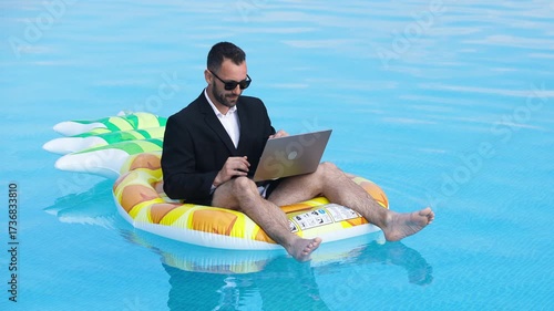 A young freelance businessman works on a laptop while sitting on an inflatable trampoline in a pool. Concepts of freelancing, remote work, and summer lifestyle.