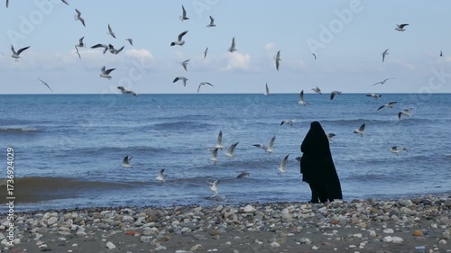 Wallpaper Mural A Muslim woman in black chador stands on the rocky shore, feeding seagulls as they circle above the waves. A poetic and cinematic seascape blending culture, faith, and nature’s freedom. Torontodigital.ca