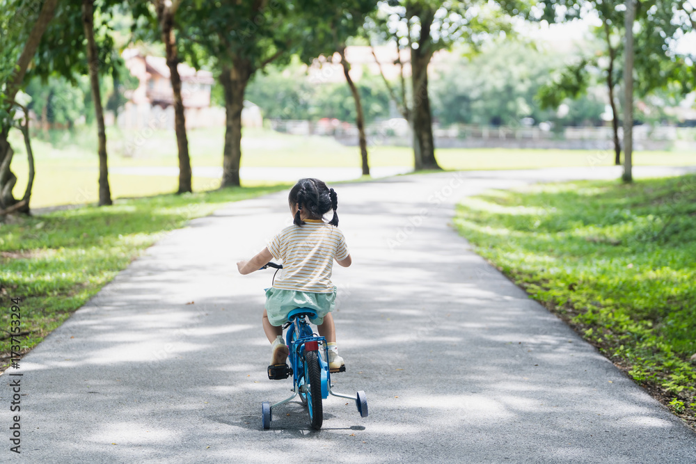Obraz premium Young child riding tricycle on pathway in serene park surrounded by trees, sunlight filtering through leaves, joyful outdoor experience, childhood adventure