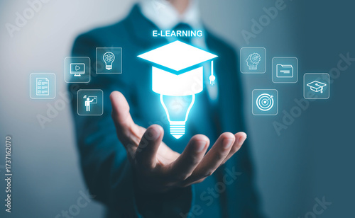 Businessman holding virtual e-learning icon with graduation cap and light bulb, symbolizing online education, digital training, remote study, knowledge innovation, and professional skill development.