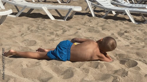 A young boy in blue swim trunks relaxes on a sandy beach, lying prone with a yellow shovel nearby, observing his surroundings on a bright day.