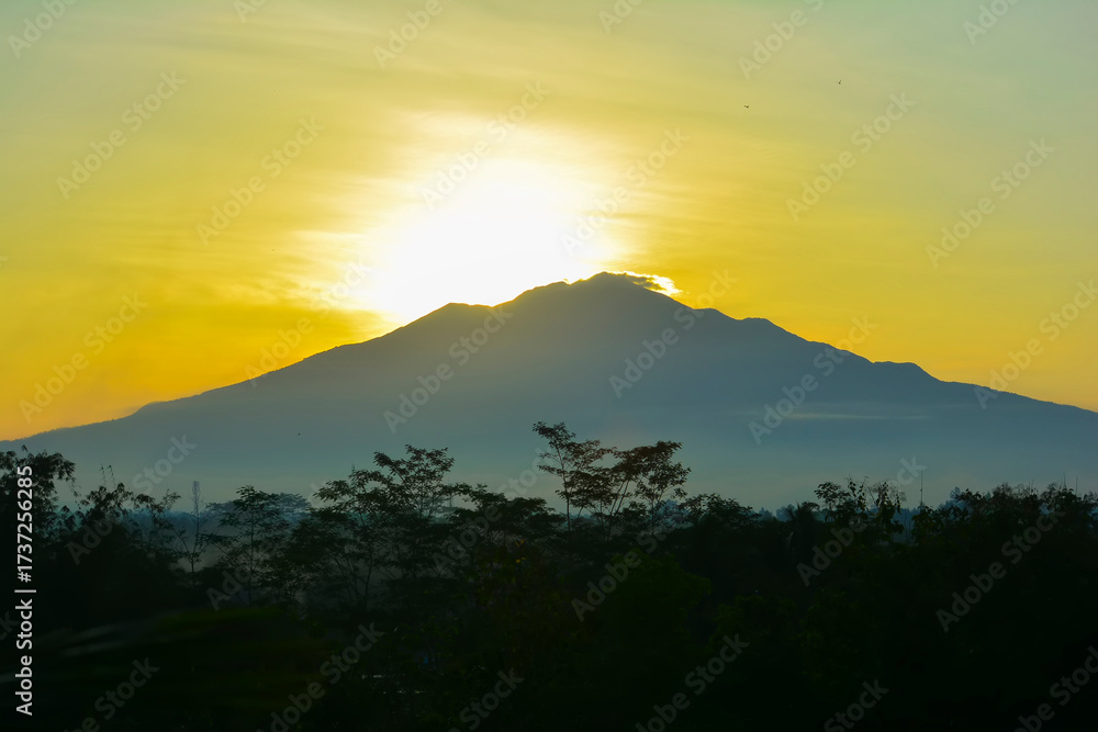Obraz premium Dramatic sunrise over Mount Merbabu in Central Java, Indonesia, with the golden morning light creating a stunning silhouette of the mountain and the surrounding trees. The scene radiates beauty