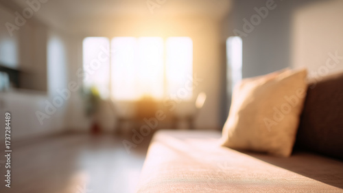 Softly lit blurred open plan home room with sunlight streaming through large windows, cozy sofa and neutral decor create warm, inviting atmosphere in modern living space