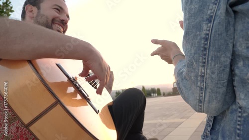Young man playing spanish acoustic guitar at sunset. A woman is clapping her hands to the rhythm of the music. Couple enjoying a romantic moment