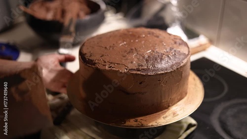 A homemade chocolate cake on a golden board being smoothed with a scraper, as hands carefully even out the creamy frosting for a perfect finish