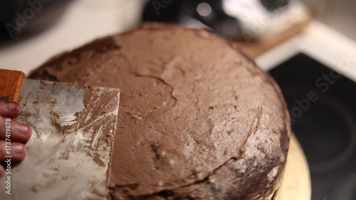 A homemade chocolate cake on a golden board being smoothed with a scraper, as hands carefully even out the creamy frosting for a perfect finish
