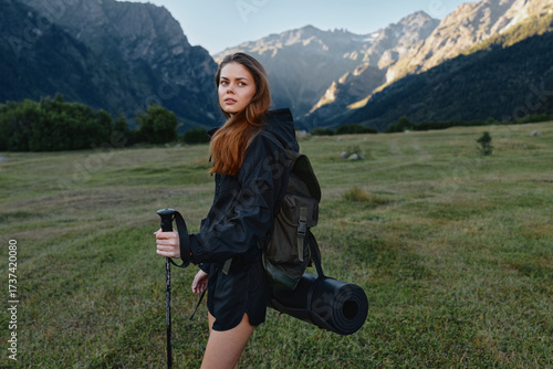 Obraz na plátně A confident hiker stands in a grassy valley with rugged mountains behind, carrying a backpack and a rolled mat