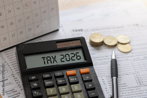 Obraz na płótnie Annual tax planning concept with calculator displaying TAX 2026, stacks of coins, a calendar, and a pen on a wooden desk