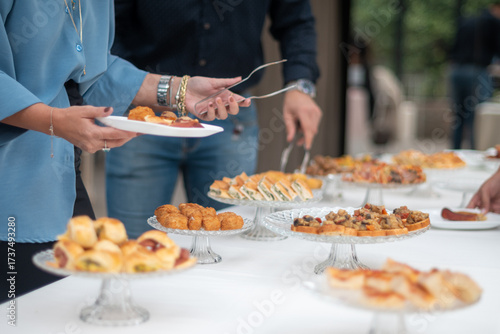 People serving themselves delicious appetizers and finger foods from a buffet table at an outdoor gathering or event showcasing a variety of tasty treats laid out on elegant display stands for guests
