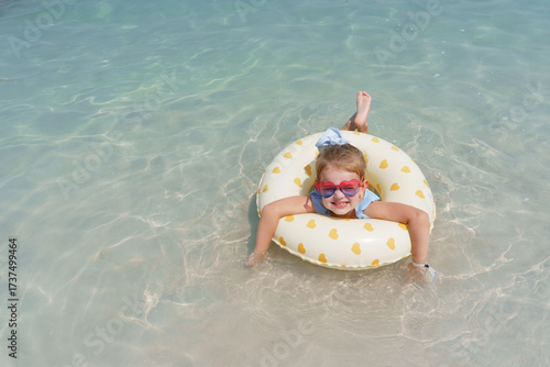 A joyful little girl with heart-shaped sunglasses smiles brightly while floating on a yellow heart-patterned inflatable ring in the clear, sun-dappled turquoise water of a tropical beach