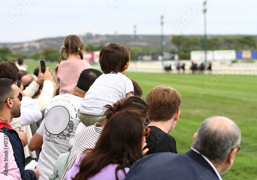 Espectadores y público con adultos y niños a hombros en caballito animando y personas atentas a carrera de caballos en pista de hierba del hipódromo