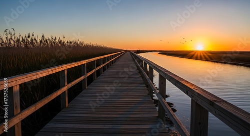 Wooden boardwalk stretching into the distance alongside a tranquil river at sunset, with golden light reflecting on the water and reeds lining the banks.