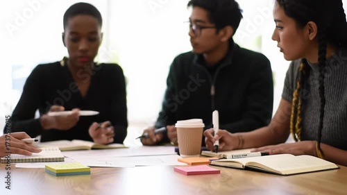 Tight video of diverse young multi-ethnic creatives collaborating around a light wooden table, gesturing at sketches/sticky notes, with soft natural light for an energetic innovation concept