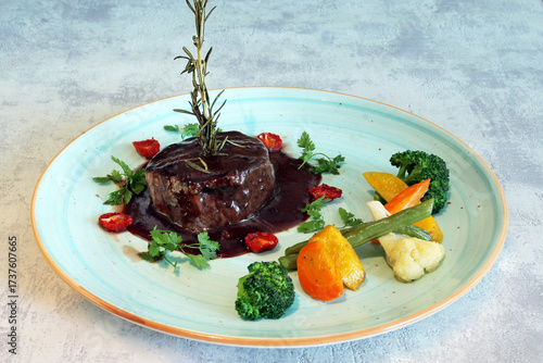 Piece of roasted beef with wine gravy and vegetables displayed on a blue plate, marble background (studio photo)