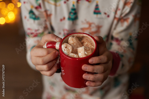 The hands of a boy in pajamas holding a mug of cocoa and marshmallows