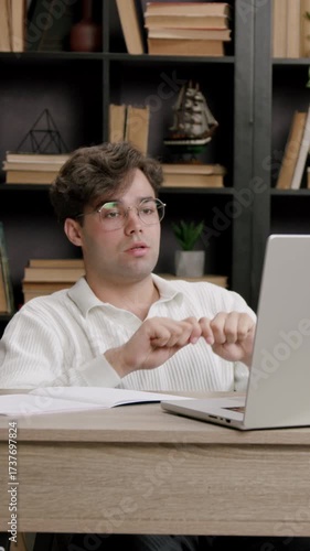 Angry boy with glasses, nervous about the large volume of lessons and projects for college. Student preparing for lessons and exams at home in a modern office with a library in the background