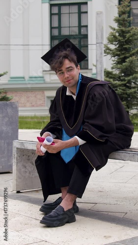 Graduate student in academic cap and gown rests happily and proudly holding his diploma on a bench near the campus. The boy thinks that all the effort was worth it