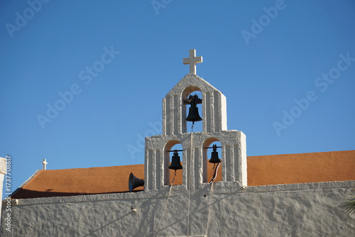 Campanile bianco con 3 campane e una croce, con tetto rosso sullo sfondo. Particolare del monastero di Odigitria sud isola di Creta