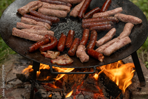 Rustic outdoor barbecue featuring a variety of sausages and meat cuts grilling on a large cast iron disc over an open wood fire. The image captures the sizzling, smoky essence of traditional cooking.