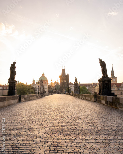 Stunning sunrise over Prague with warm golden light illuminating historic architecture, cobblestone streets, and iconic city landmarks in high-quality detail.