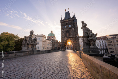 Stunning sunrise over Prague with warm golden light illuminating historic architecture, cobblestone streets, and iconic city landmarks in high-quality detail.