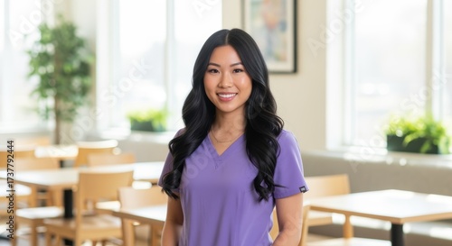 A young Asian woman in purple medical scrubs smiles warmly in a bright, modern healthcare facility. Natural light fills the space. Perfect for medical or nursing profession concepts.
