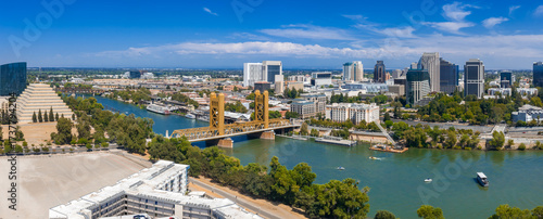 Aerial view of Sacramento, California, featuring the golden Tower Bridge, Sacramento River, downtown skyline, and ziggurat shaped building under a clear sky.