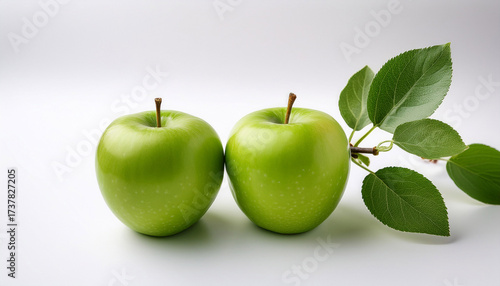 Two Bright Green Apples Rest On A White Surface Accompanied By A Small Branch With Several Green Leaves The Apples Are Positioned Slightly Off Center Creating A Balanced Composition