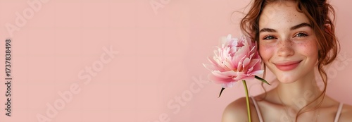 A young woman with freckles joyfully holds a pink flower close to her face. Her warm smile and natural beauty create a charming atmosphere against a soft pastel backdrop