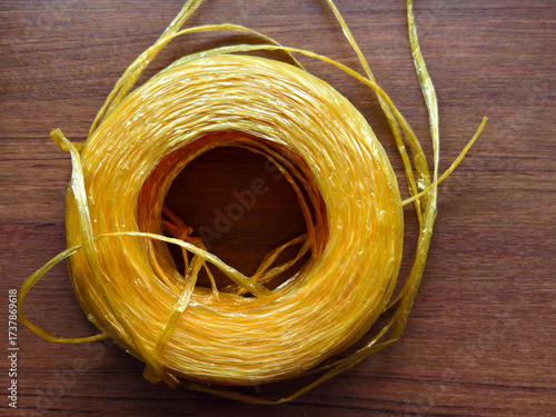 Close-up of yellow raffia plastic rope roll on wooden surface. Strong, durable, and flexible cord used for packaging, shipping, agriculture, DIY, and craft projects.