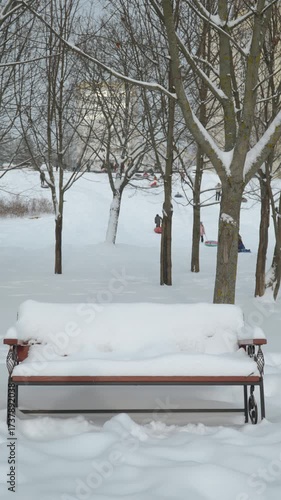 Vertical video of snow covered bench in city park. In distance behind trees, blurred people and children are sliding down hill on snow tube. Natural backdrop of snowy day after snowstorm.