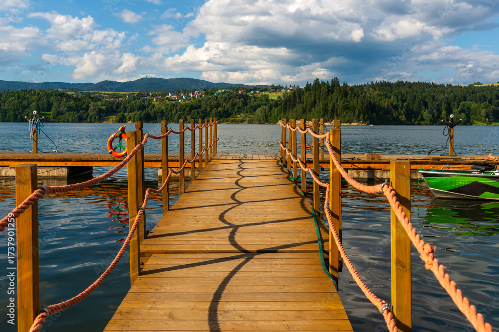 Naklejka premium A wooden pier with orange rope railings casts rhythmic curved shadows on the water surface. Clear sky with fluffy clouds and forested hills create a calm, inviting summer lakeside scene.