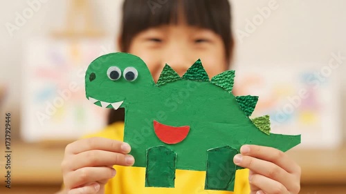 Girl holding green dinosaur craft with googly eyes paper teeth spikes and red stomach detail