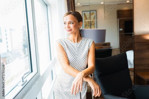 Woman standing by window in hotel room with suitcase.