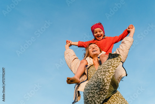 Mother carrying her daughter on her shoulder against blue sky. Smiling mom carrying happy little girl making airplane arms. Young family have fun at beach in windy winter.
