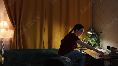 Young woman student sits indoors at desk doing homework in softly lit evening room focused on education and preparing for academic success at home. Copy space