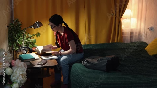 Beautiful student girl sits indoors in evening home setting at desk learning with books and laptop preparing for college exams and courses. Copy space