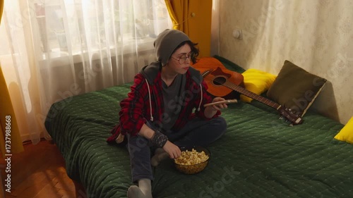 Stylish teenager in modern fashion outfit enjoys popcorn on bed while using smartphone and watching TV, concept of cozy indoor entertainment and leisure.