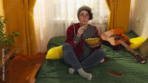 Teen girl with long brunette hair in glasses and stylish clothes enjoys popcorn while sitting on bed and watching television, youth leisure and comfort. Mid shot