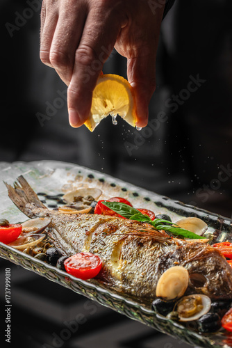 Close-up of a chef squeezing lemon juice on baked dorado fish served with cherry tomatoes, basil, olives, and clams. Gourmet Mediterranean seafood dish concept