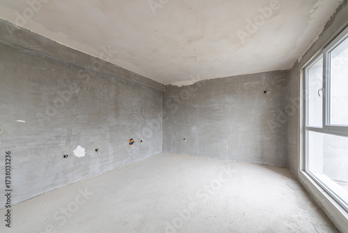 Interior of an empty room under construction in a new apartment. Unfinished space with raw concrete walls, floor, and a large window. Renovation and real estate concept.