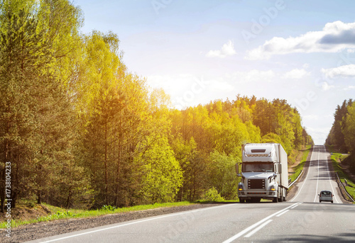 American semi-trailer truck transports cargo along country road uphill against sun and forest in summer. Copy space for text, logistics company